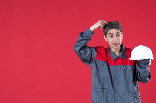 Vista frontale ravvicinata di pensare giovane lavoratore in uniforme che tiene elmetto sul muro rosso isolato