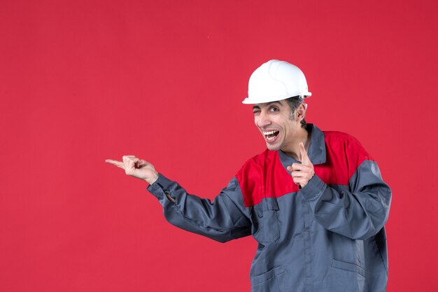 Vista frontale ravvicinata di un giovane lavoratore sorridente e divertente in uniforme con elmetto rivolto verso l'alto sul muro rosso isolato