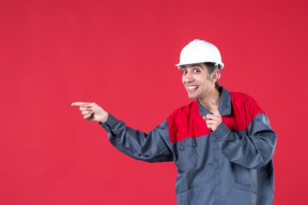 Vista frontale ravvicinata di un giovane lavoratore sorridente felice in uniforme con elmetto rivolto verso l'alto sul muro rosso isolato