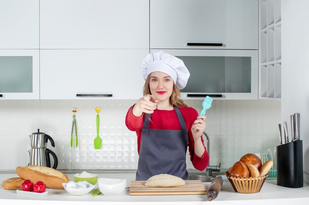 Vista frontale soddisfatta donna bionda con cappello da cuoco e grembiule che punta alla telecamera in cucina