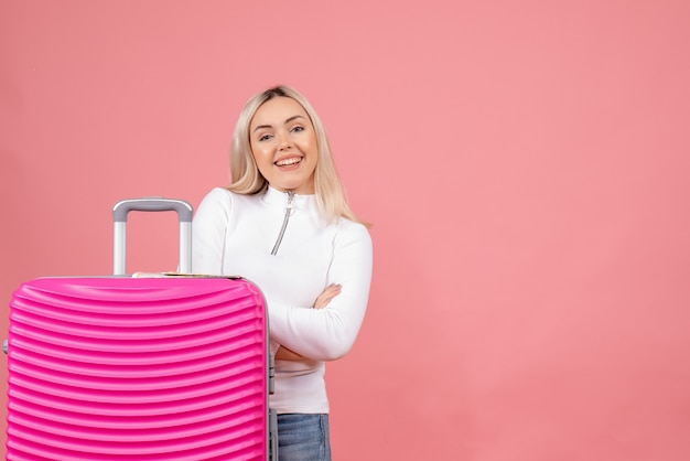 Vista frontale sorridente giovane signora in piedi dietro la valigia rosa