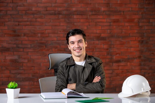 Vista frontale sorridente ingegnere maschio seduto dietro il suo posto di lavoro lavoro affari appaltatore agenda costruttore aziendale documenti del piano di proprietà