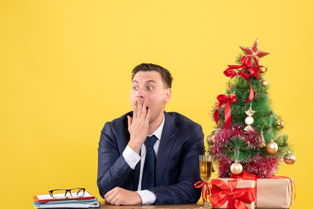 Vista frontale stupito uomo seduto al tavolo vicino albero di Natale e regali su sfondo giallo spazio libero