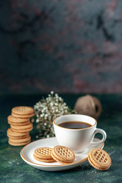 Vista frontale tazza di tè con biscotti dolci nel piatto bianco sulla parete scura colore del pane cerimonia colazione mattina bicchiere bevanda zucchero foto