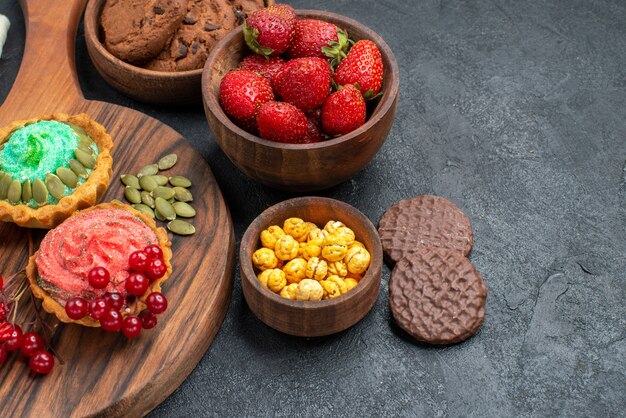 Vista frontale torte cremose con frutta e biscotti su sfondo scuro