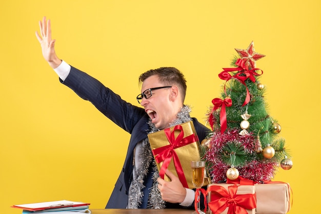 Vista frontale uomo arrabbiato aprendo la mano seduto al tavolo vicino all'albero di Natale e presenta su sfondo giallo