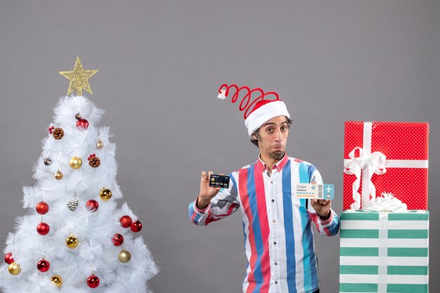Vista frontale uomo confuso che tiene carta e biglietto di viaggio intorno all'albero di Natale e regali