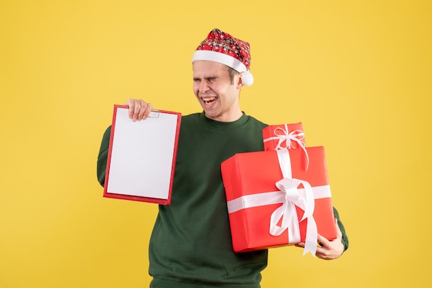 Vista frontale uomo euforico con maglione verde che tiene grande regalo e appunti in piedi sul giallo