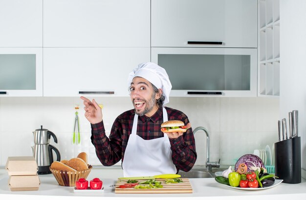 Vista frontale uomo sorpreso che regge un hamburger in piedi dietro il tavolo della cucina