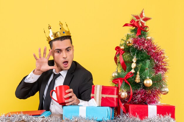 Vista frontale uomo sorpreso con corona guardando tazza vicino albero di Natale e regali