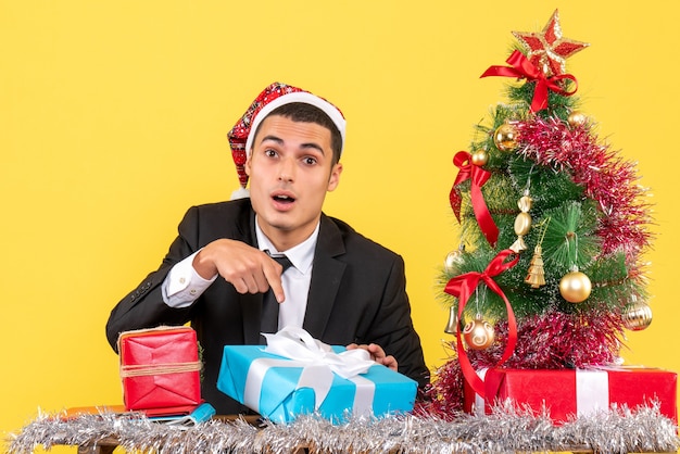 Vista frontale uomo sorpreso con il cappello della santa seduto al tavolo che mostra il suo albero di natale e regali attuali