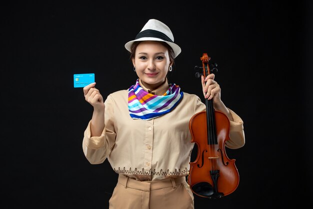 Vista frontale violinista femminile che tiene violino e carta di credito sulla parete scura melodia strumento musica emozione concerto spettacolo performance woman