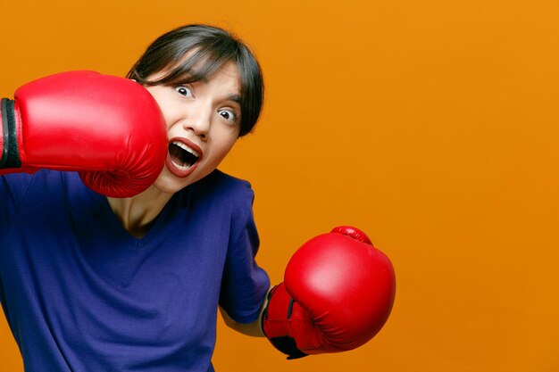 Vista ingrandita della giovane donna sportiva spaventata che indossa maglietta e guanti da boxe guardando la telecamera che si colpisce in faccia isolata su sfondo arancione