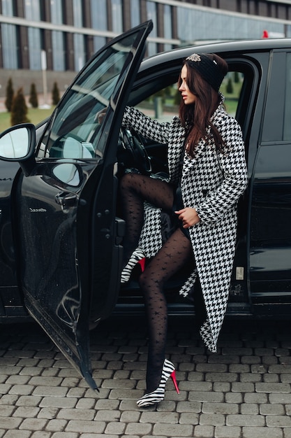 Vista laterale a figura intera di una donna bruna alla moda con un elegante cappotto stampato a zampe di gallina e tacchi zebrati seduta nella sua auto al volante...