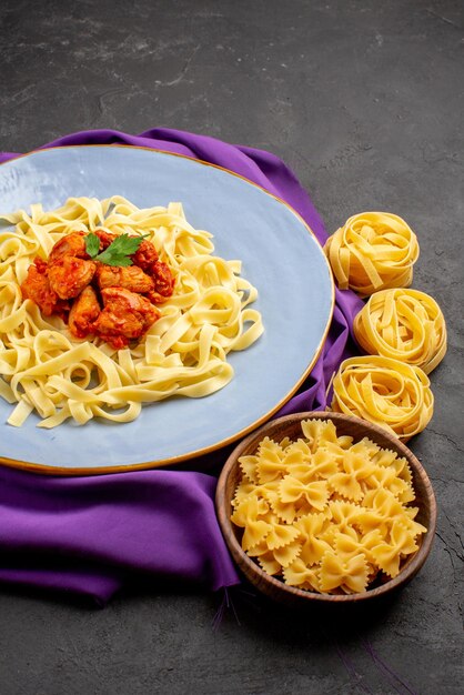 Vista laterale da lontano carne e pasta carne e sugo sulla tovaglia viola e ciotole di pasta sul tavolo scuro