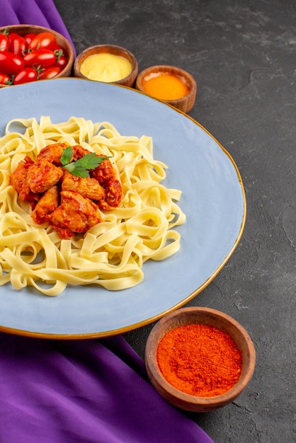 Vista laterale da lontano pasta e salse piatto di pasta con sugo e carne accanto alle ciotole di pomodori e salse sulla tovaglia viola sul tavolo scuro