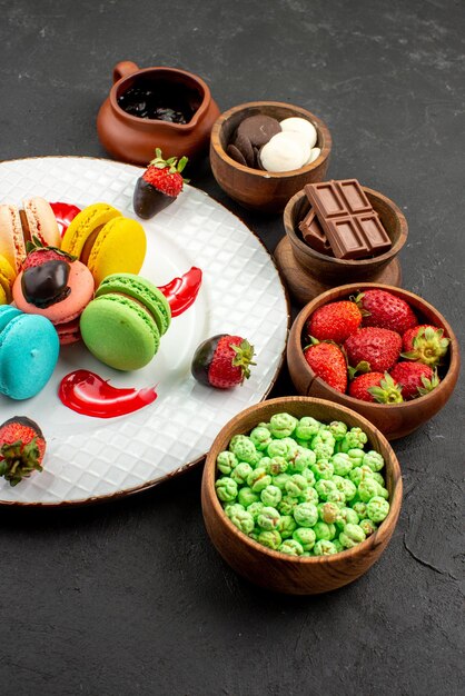 Vista laterale da lontano piatto di dolci di amaretti e ciotole di fragole al cioccolato caramelle verdi e crema al cioccolato sul tavolo