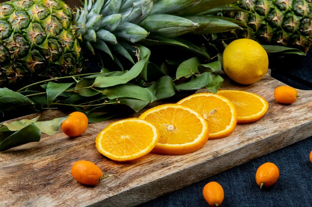 Vista laterale degli agrumi come arancia e kumquat con le foglie sul tagliere con gli ananas sul fondo del panno dei jeans