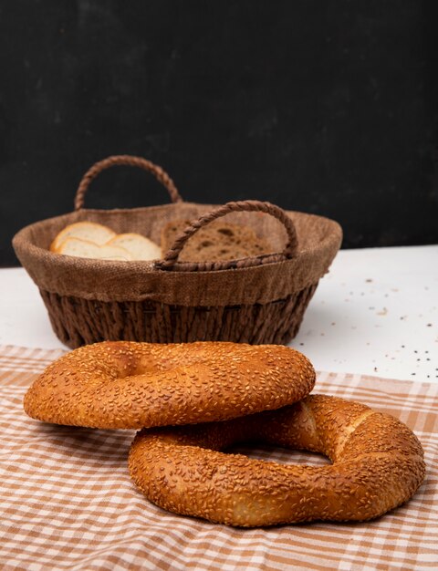 Vista laterale dei bagel sul panno con il canestro delle fette bianche e del pane di segale su superficie bianca e fondo nero con lo spazio della copia