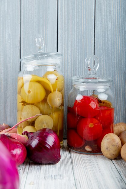 Vista laterale dei barattoli di vetro con le patate acide delle cipolle e dei pomodori su superficie e su fondo di legno con lo spazio della copia