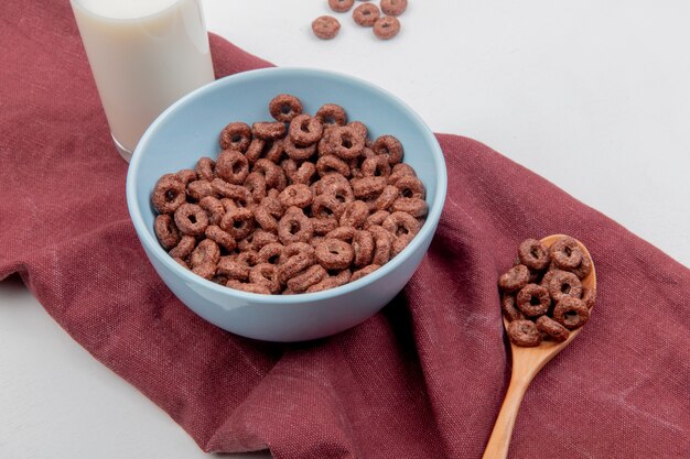 Vista laterale dei cereali in una ciotola e in un cucchiaio di legno con latte sul panno bordo e superficie bianca