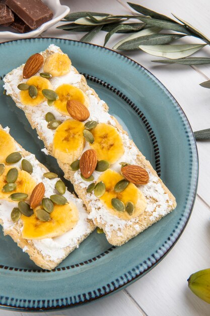 Vista laterale dei cracker croccanti con crema di formaggio, fette di banana, mandorle e semi di zucca su un piatto su legno bianco