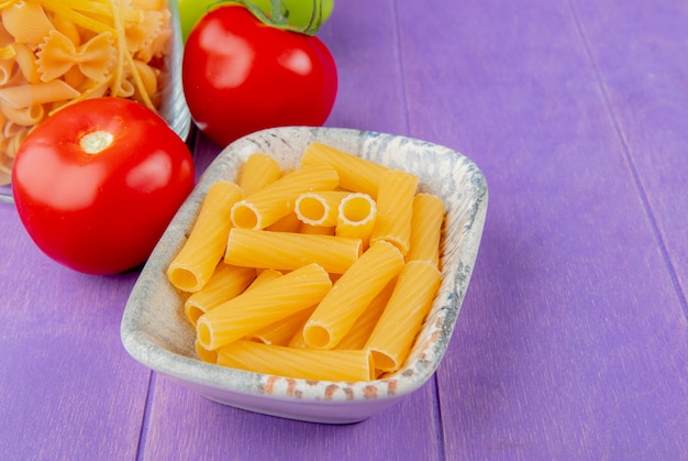 Vista laterale dei maccheroni del penne e di altri tipi in ciotole e pomodori sulla tavola porpora con lo spazio della copia