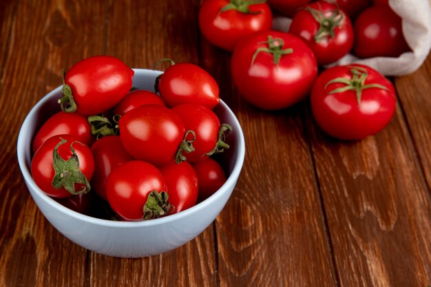 Vista laterale dei pomodori in ciotola con altri che si rovesciano dal sacco sulla tavola di legno