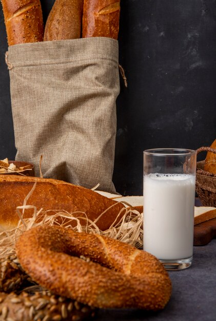 Vista laterale del bagel e del bicchiere di latte con il sacco delle baguette sulla superficie marrone rossiccio e sul fondo nero con lo spazio della copia