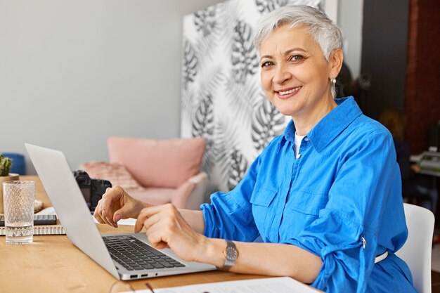 Vista laterale del blogger femminile maturo di successo seduto in ufficio a casa e laptop aperto, digitazione, scrittura di un nuovo articolo sulla psicologia con un ampio sorriso sicuro