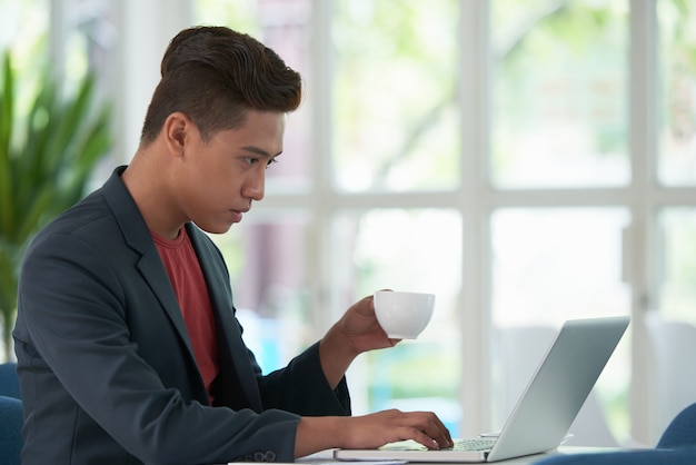 Vista laterale del caffè sorseggiante del tipo asiatico e lavorare al computer portatile