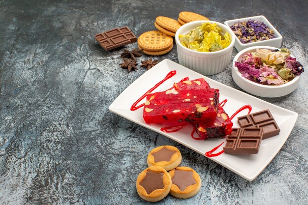 Vista laterale del cioccolato su piatto bianco con ciotole di fiori secchi e biscotti su fondo grigio