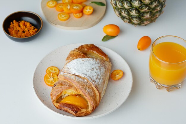 Vista laterale del croissant con fette di kumquat nel piatto e scorza d'arancia di ananas con succo d'arancia su sfondo bianco