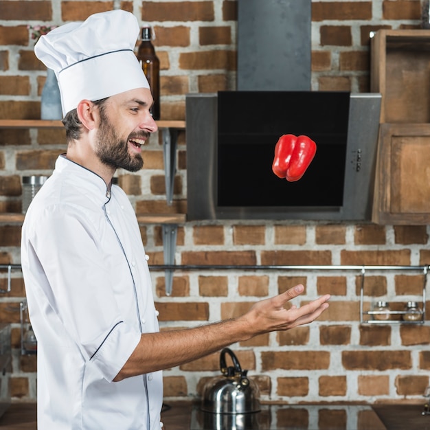 Vista laterale del cuoco unico maschio che getta peperone dolce rosso nell&#39;aria