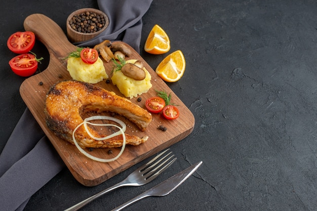 Vista laterale del delizioso pasto di pesce fritto con funghi verdure formaggio su tavola di legno fette di limone pepe su posate asciugamano di colore scuro impostato su superficie nera in difficoltà