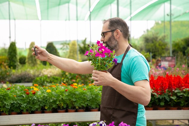 Vista laterale del giardiniere dai capelli grigi prendendo selfie con la petunia