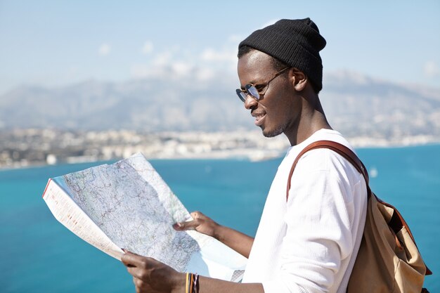 Vista laterale del giovane autostoppista sorridente che studia mappa cartacea nelle sue mani