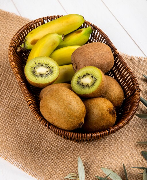 Vista laterale del kiwi e della banana freschi in un canestro di vimini su tela di sacco su bianco