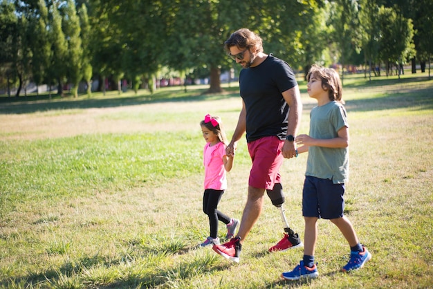 Vista laterale del padre con disabilità che cammina con i bambini. Uomo dai capelli ondulati con gamba meccanica che cammina nel parco con un ragazzino e una ragazza. Disabilità, famiglia, concetto di amore