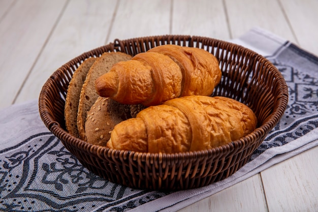 Vista laterale del pane come croissant e fette di pane pannocchia marrone seminate nel cestino sul panno su fondo di legno