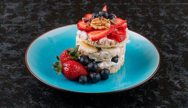 Vista laterale del pane croccante saporito con le fragole e i dadi maturi dei mirtilli con panna acida su un piatto ceramico su superficie rustica di legno