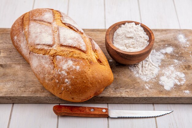 Vista laterale del pane di grano fatto in casa con una ciotola di farina sul tagliere e coltello su fondo in legno