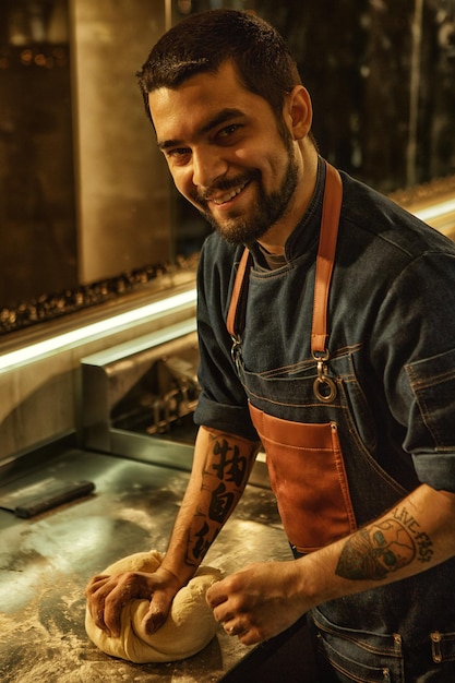 Vista laterale del panettiere maschio sorridente e positivo che prepara e rotola la pasta sul tavolo di metallo ricoperto di farina bell'uomo con barba e tatuaggi sulle mani che indossano un grembiule di jeans