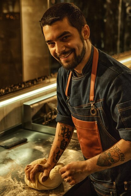 Vista laterale del panettiere maschio sorridente e positivo che prepara e rotola la pasta sul tavolo di metallo ricoperto di farina bell'uomo con barba e tatuaggi sulle mani che indossano un grembiule di jeans