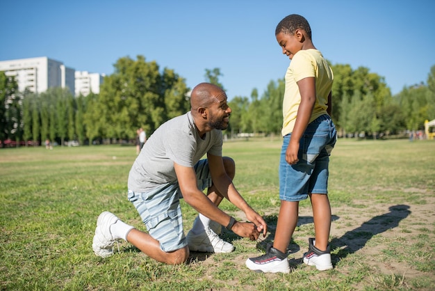 Vista laterale del papà africano e di suo figlio in piedi sul campo. Uomo sorridente in piedi su un ginocchio sull'erba che si allaccia i lacci delle scarpe su scarpe da ginnastica per bambini che si guardano l'un l'altro. Cura dei genitori e concetto di attività sportiva