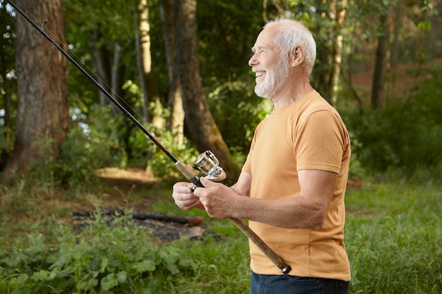 Vista laterale del pensionato maschio europeo bello che cattura pesci contro gli alberi di pino, tirando l'asta con cattura fuori dall'acqua, sorridendo felicemente, godendo di hobby all'aperto attivo nella natura selvaggia