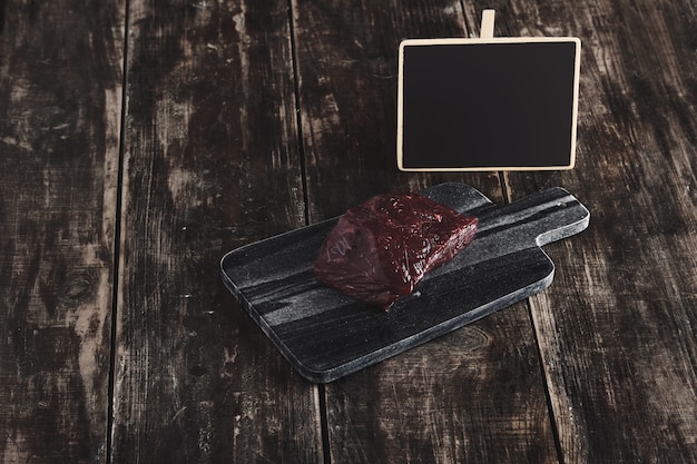 Vista laterale del pezzo grezzo di lusso di bistecca di carne di balena sul banco di taglio di pietra di marmo nero e tavolo in legno vintage invecchiato e cartellino del prezzo del bordo di gesso