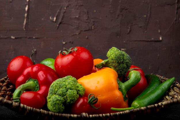 Vista laterale del piatto del canestro pieno delle verdure come broccoli e pomodori dei peperoni sulla superficie nera e sulla superficie marrone rossiccio
