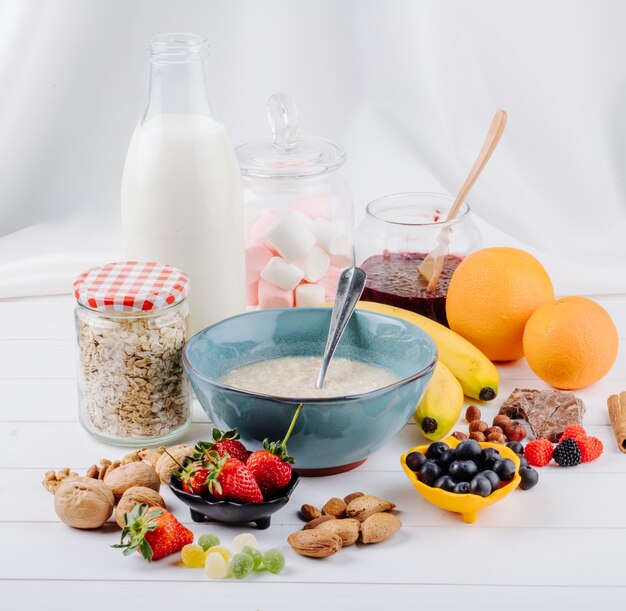 Vista laterale del porridge della farina d'avena in una ciotola e bacche fresche arance e noci della banana sulla tavola rustica bianca