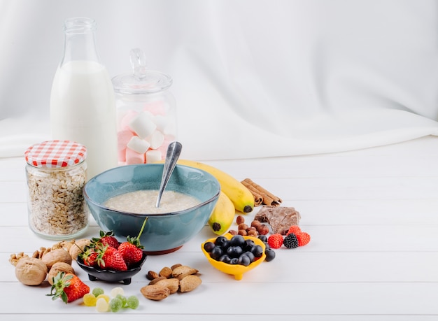 Vista laterale del porridge di farina d'avena in una ciotola e bacche fresche banana e noci su fondo rustico bianco